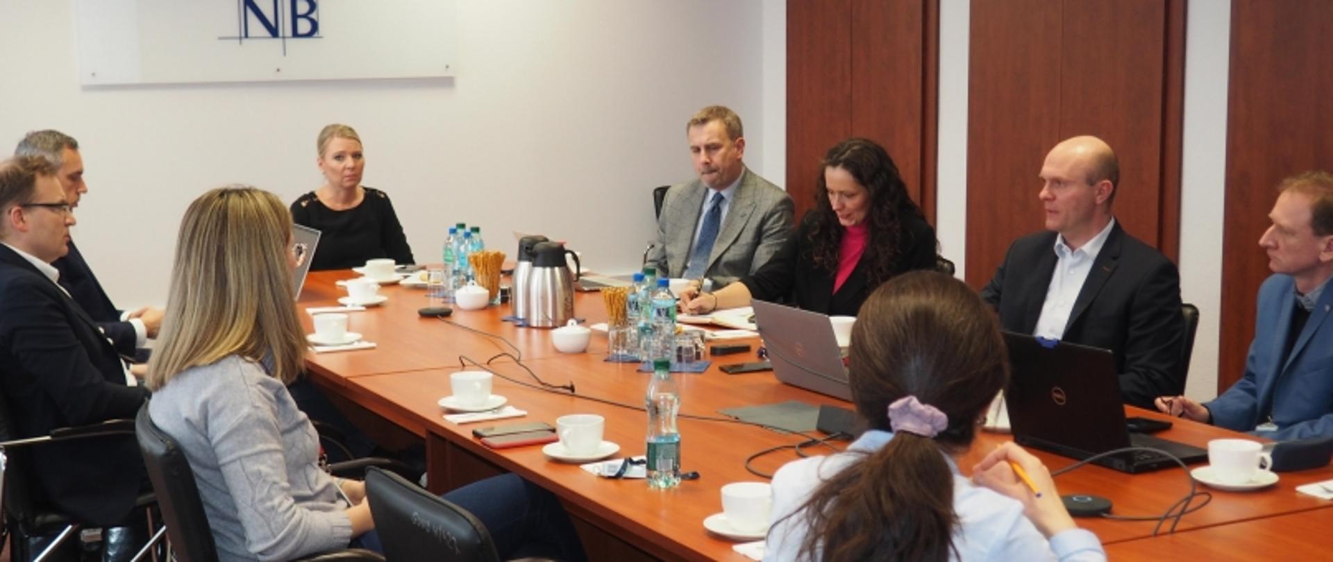 Grupa ośmiu osób przy stole konferencyjnym z laptopami, notatnikami i napojami; w tle znak z literami „NB”.