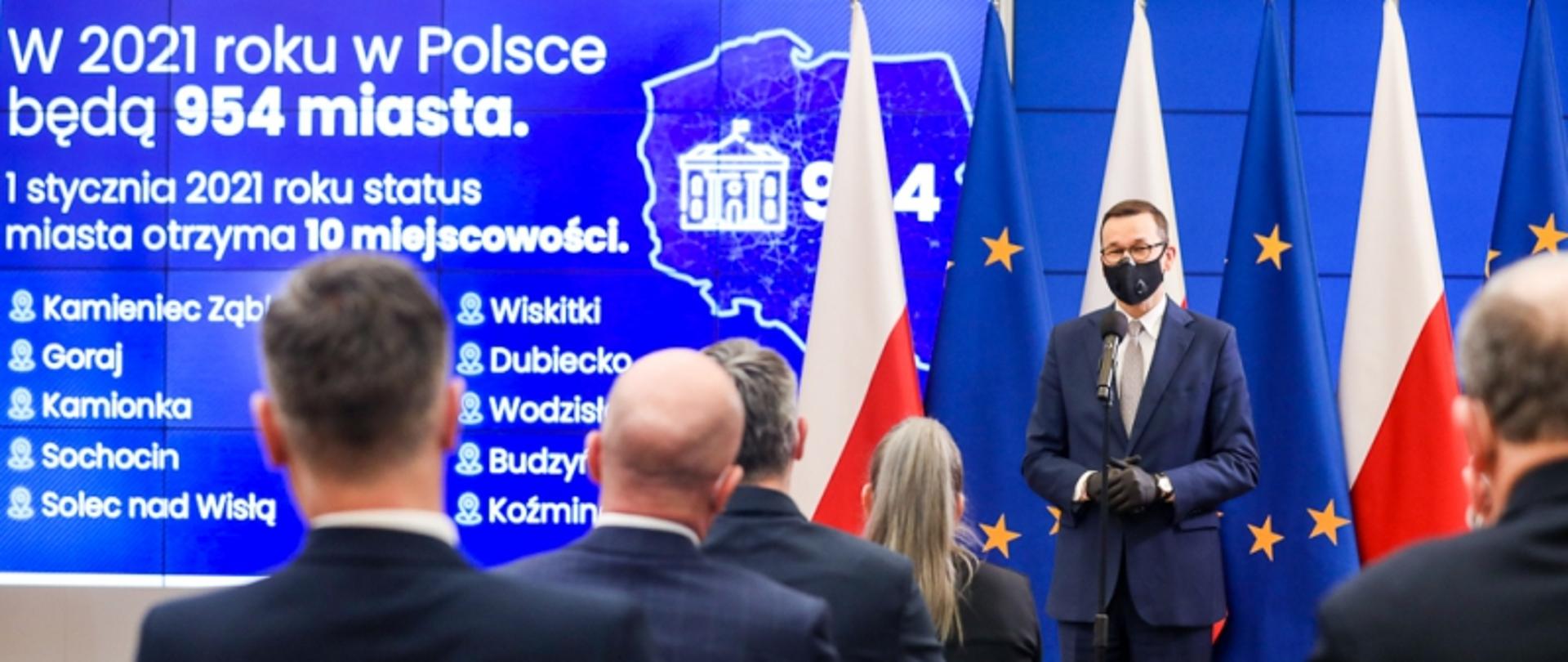 Zdjęcie przedstawia uroczystość przekazania aktów nadania statusu miasta kolejnym 10 miejscowościom. Na pierwszym planie znajduje się pięć osób, odwróconych tyłem do fotografa, którzy siedzą w fotelach. Przed nimi znajduje się mężczyzna ubrany w granatowy garnitur, koszulę i krawat. W tle znajdują się biało-czerwone flagi oraz flagi Unii Europejskiej oraz telebim. 