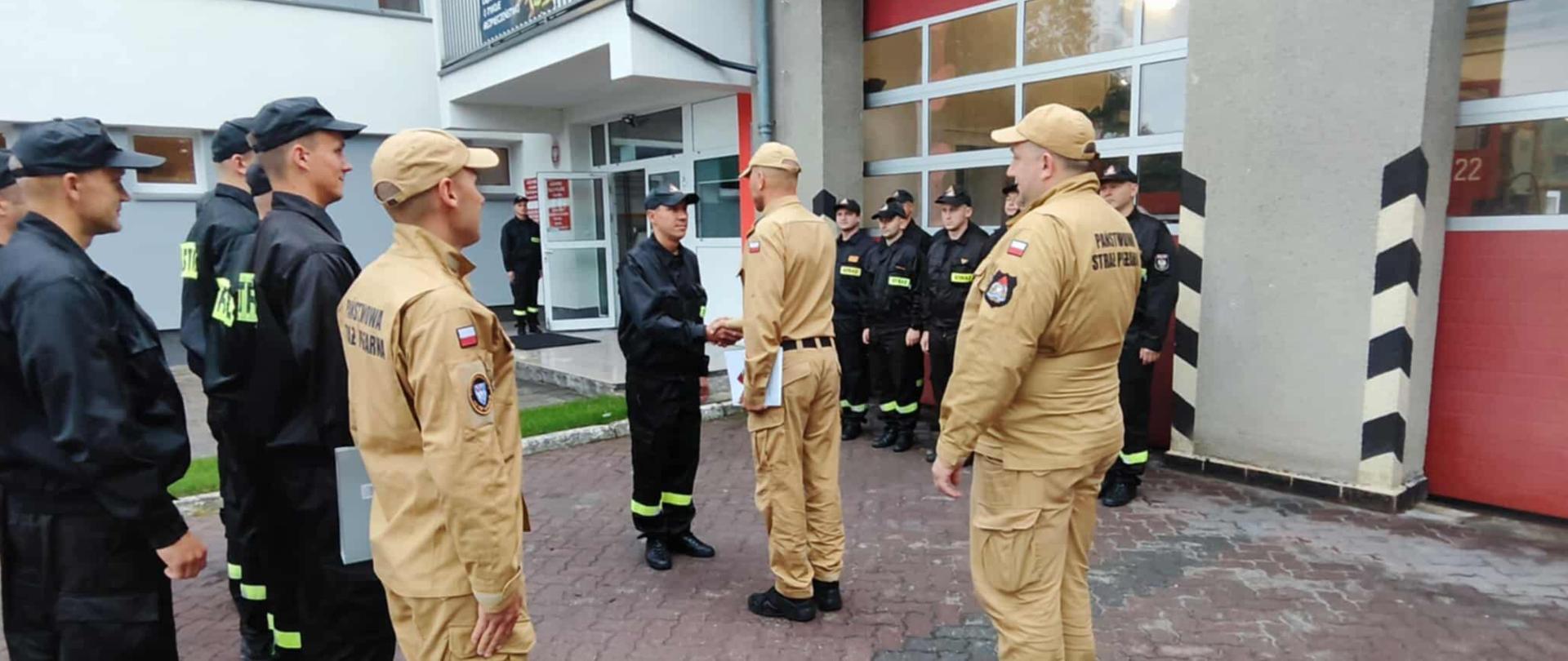Na placu przed garażem strażnicy odbywa się uroczyste powitanie – funkcjonariusz w czarnym mundurze podaje rękę strażakowi w piaskowym mundurze. Obok stoją pozostali strażacy, tworząc dwa szeregi. W tle czerwona brama garażowa