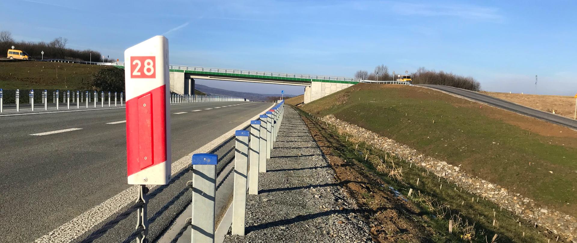 Zdjęcie przedstawia fragment nowej drogi z barierami ochronnymi po lewej stronie. Na pierwszym planie stoi słupek z oznaczeniem „28”. W tle widać zielony wiadukt nad drogą oraz łagodne skarpy z trawą po obu stronach. Niebo jest jasne i bezchmurne.