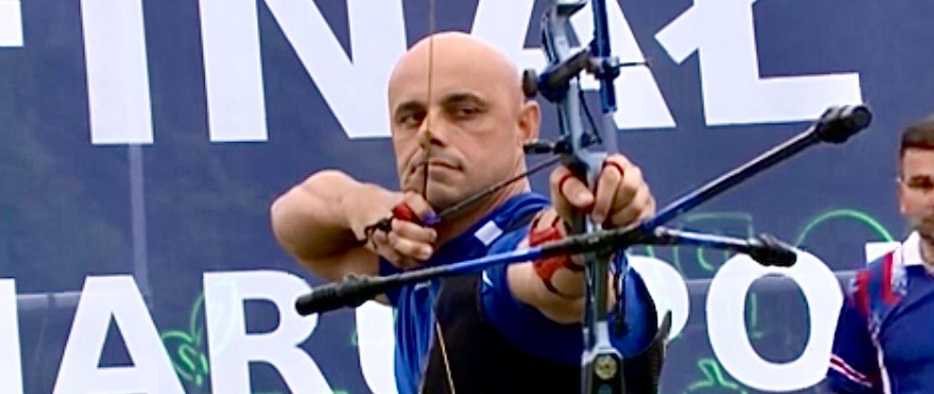 Zawodnik stoi przodem, trzyma łuk, przygotowany do strzału, w tle niebieski baner, na dole tablica z wynikami