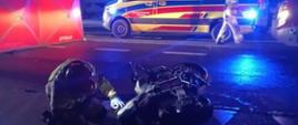 Zniszczony motocykl po wypadku drogowym. W tle karetka oraz funkcjonariusz PSP.