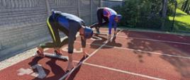 Zdjęcie zrobione z boku i z góry, przedstawiające dwóch strażaków w pozycji startowej na bocznych torach bieżni lekkoatletycznej o czerwonej, chropowatej nawierzchni. Na torze po lewej stronie (oznaczonym namalowaną białą cyfrą 1) klęczy w blokach startowych zawodnik w ciemnoniebiesko-czarnym stroju sportowym z żółtymi elementami oraz w ciemnoniebieskim kasku. Jego dłonie opierają się o linię startową, a sylwetka rzuca wyraźny cień przed nim. Na torze obok niego, nieco z tyłu, w identycznej pozycji startowej znajduje się drugi zawodnik w niebiesko-biało-czerwonej koszulce i czarnych getrach.
W tle, po lewej stronie, biegnie wysoki, szary płot z betonowych prefabrykatów. Za nim widoczne są drzewa oraz budynek parterowego warsztatu samochodowego z niebieskimi tablicami informacyjnymi (m.in. z logo marki Elf). Nad całą sceną panuje bezchmurne, słoneczne niebo. W prawym dolnym rogu zdjęcia umieszczono mały podpis: „st. str. Agnieszka Wardecka | Państwowa Straż Pożarna”.
