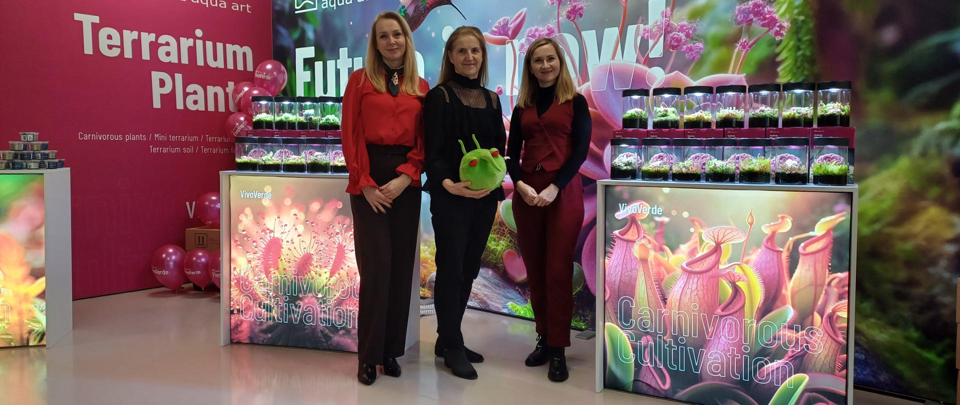 Na zdjęciu widać stoisko wystawowe. W tle znajdują się duże plansze z napisami „Terrarium Plants” oraz „Future is now” z grafikami roślin i kolibrem. Przy planszach widać balony w kolorze różowym. Na ladach ekspozycyjnych ustawiono przezroczyste, cylindryczne pojemniki (terraria) z roślinami w środku. Fronty lad zdobią zdjęcia roślin wodnych i owadożernych. W centralnej części zdjęcia trzy uśmiechające się kobiety, jedna z nich trzyma zieloną kulistą, pluszową maskotkę Pesty.