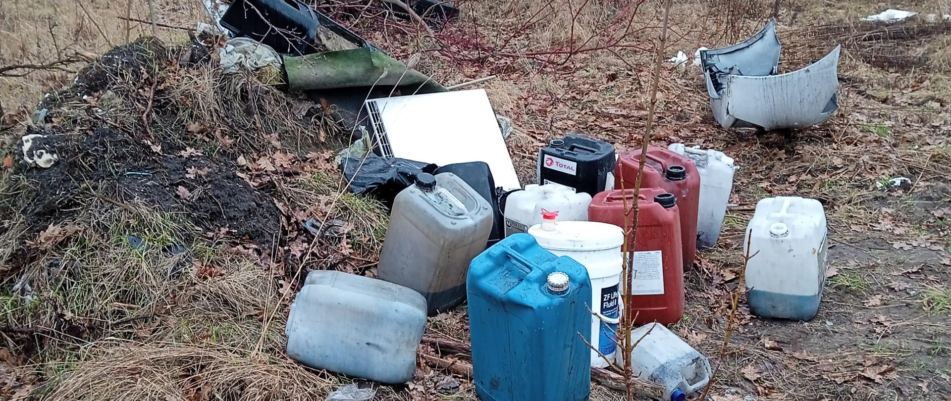 Kilkanaście plastikowych pojemników, w większości 20 litrowych, stoi na nieutwardzonym podłożu. W tle znajdują się wątłe drzewa.