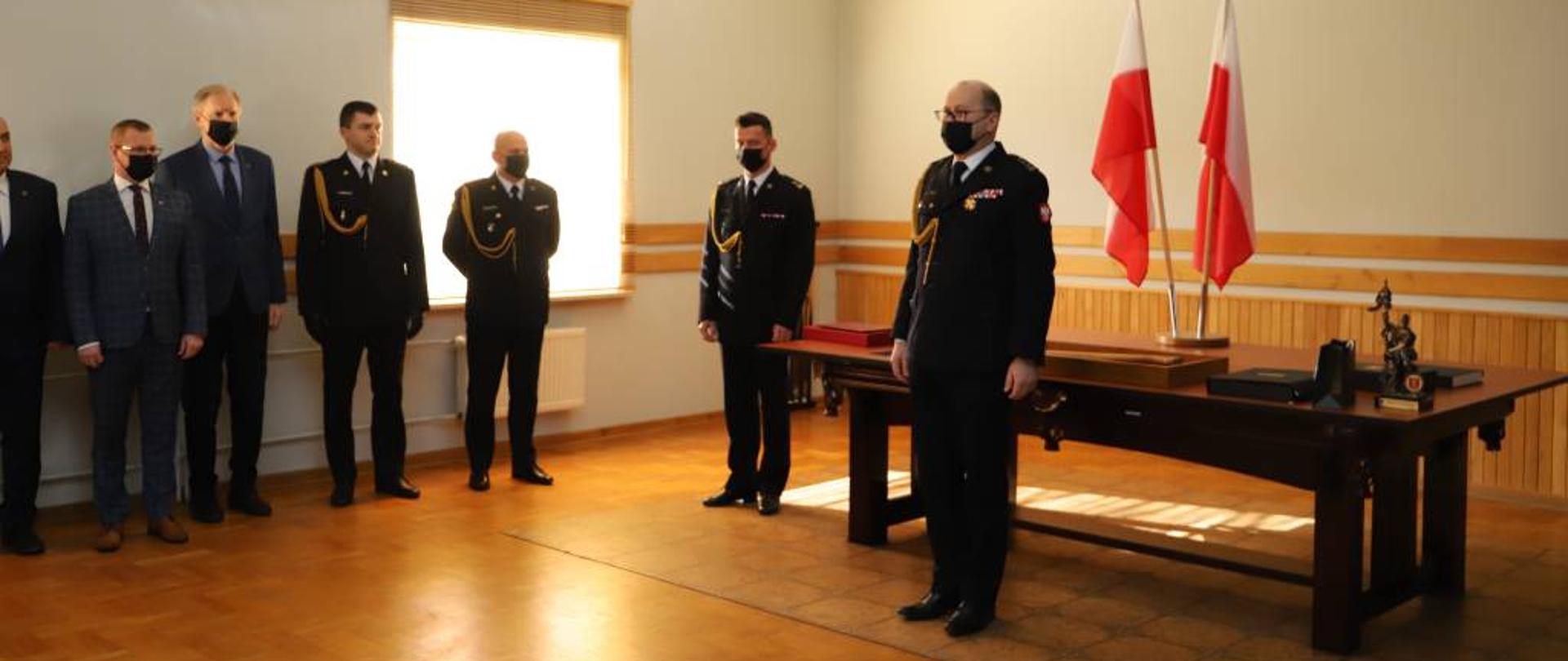 Zdjęcie przedstawia starszego brygadiera Krzysztofa Konickiego w mundurze wyjściowym ze sznurem galowym podczas przemowy z okazji zdania obowiązków Komendanta Powiatowego Państwowej Straży Pożarnej w Zambrowie. Z lewej strony zdjęcie objęło kilku z zaproszonych gości oraz trzech funkcjonariuszy Państwowej Straży Pożarnej w mundurach wyjściowych ze sznurem galowym. Za zdającym obowiązki Komendanta Powiatowego Państwowej Straży Pożarnej w Zambrowie starszym brygadierem Krzysztofem Konickim znajduje się stół na którym leży kilka pamiątek wręczanych przez gości oraz pracowników z okazji przejścia na emeryturę. Uroczystość odbywała się w jednym z pomieszczeń Komendy Powiatowej Państwowej Straży Pożarnej w Zambrowie. 