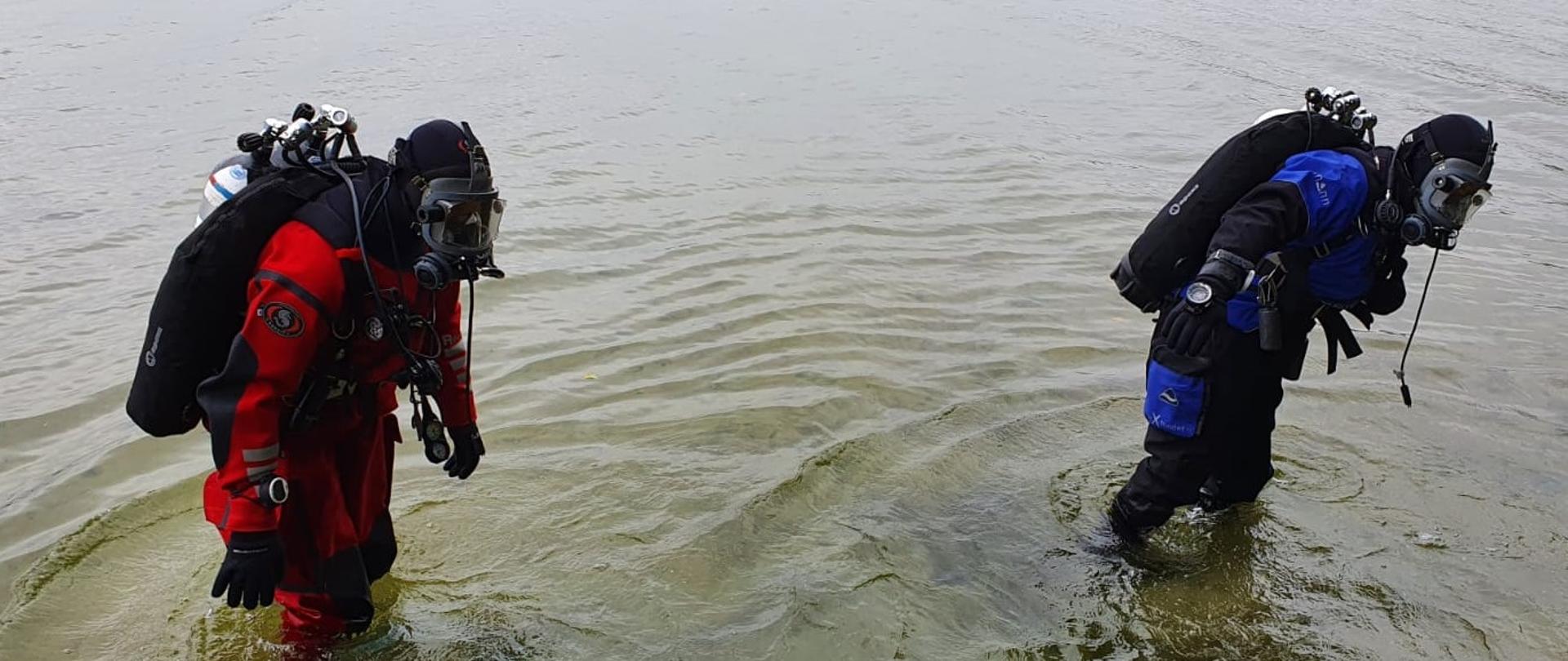 Dwóch kompletnie ubranych nurków w zestawy nurkowe wychodzących z jeziora. Jeden ma suchy skafander koloru czerwonego a drugi koloru niebieskiego. 