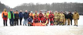 Uczestnicy ćwiczeń wodno-lodowych na grupowym zdjęciu stoją w rzędzie wraz ze sprzętem, zarówno strażacy jak i przedstawiciele firmy