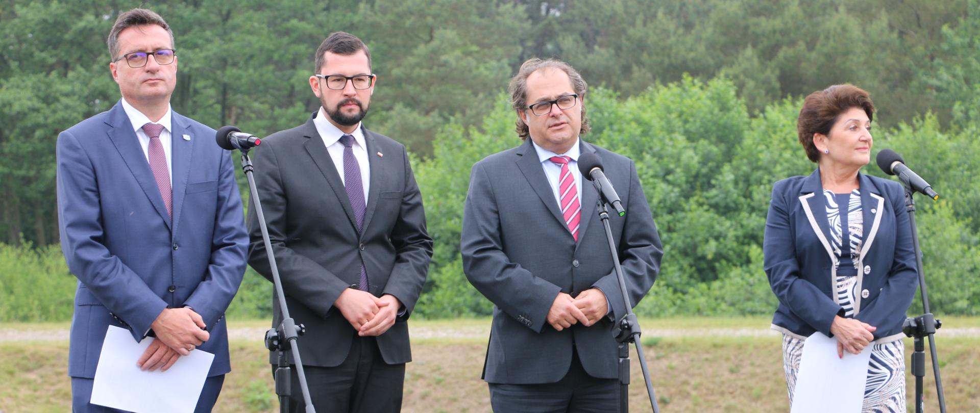 Uczestnicy konferencji stoją przy mikrofonach, w tle kanał jamneński i drzewa.