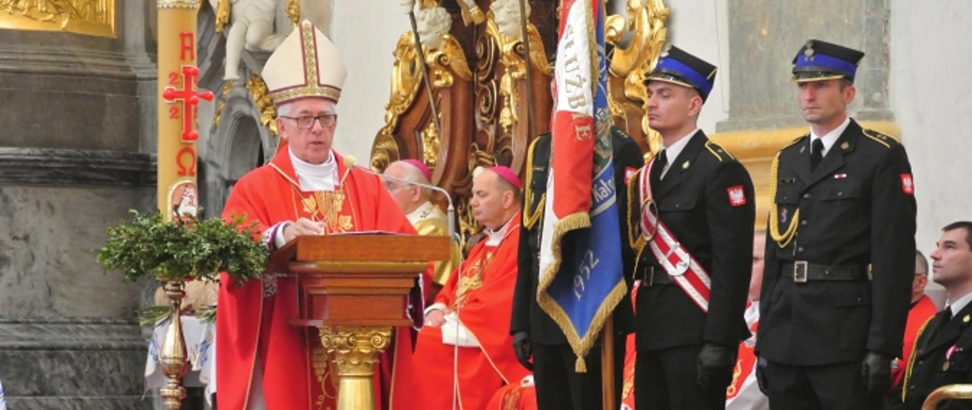 Biskup przemawia podczas mszy stojąc przy mównicy. Po prawej widoczny poczet sztandarowy (trzech strażaków). W tle siedzący księża