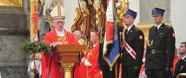 Biskup przemawia podczas mszy stojąc przy mównicy. Po prawej widoczny poczet sztandarowy (trzech strażaków). W tle siedzący księża