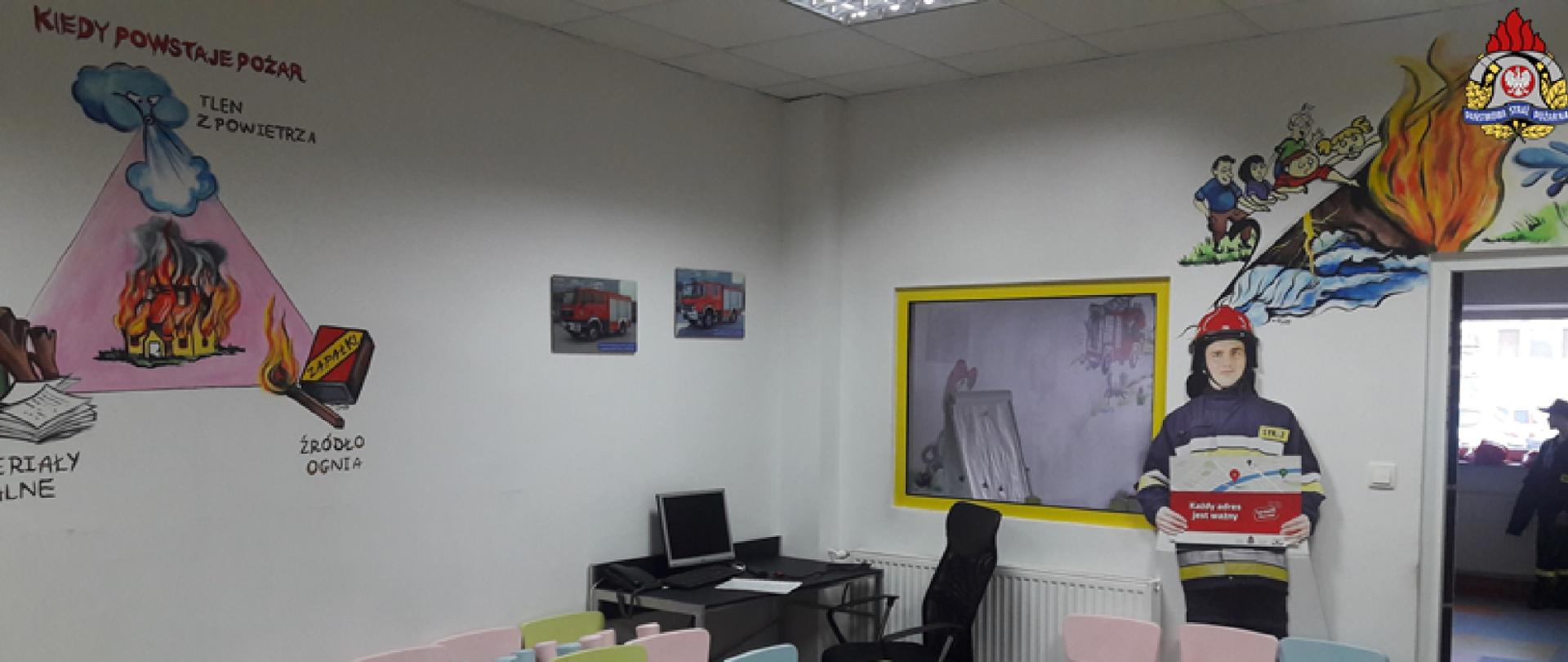 Na zdjęciu widoczna jest sala edukacyjna dla dzieci. Możemy zobaczyć poukładane kolorowe krzesła, plakat strażaka oraz stanowisko komputerowe. Ściany pomalowane na biało przyzdobione są fotografiami pojazdów pożarniczych oraz namalowany na nich jest tzw. trójkąt pożarowy (płynącą dom, tlen z powietrza, materiały palne oraz źródło ognia).