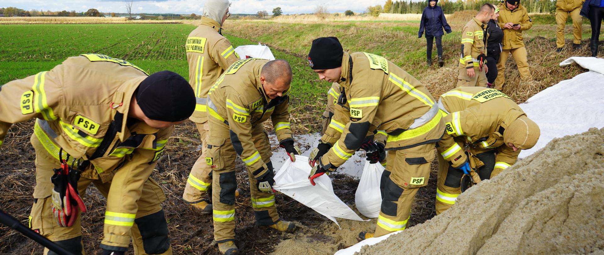 Strażacy PSP napełniają worki piaskiem, tworząc zaporę przeciwpowodziową na otwartym polu. W tle widać obserwatorów i pochmurne niebo, a cała scena ukazuje ich współpracę i zaangażowanie.