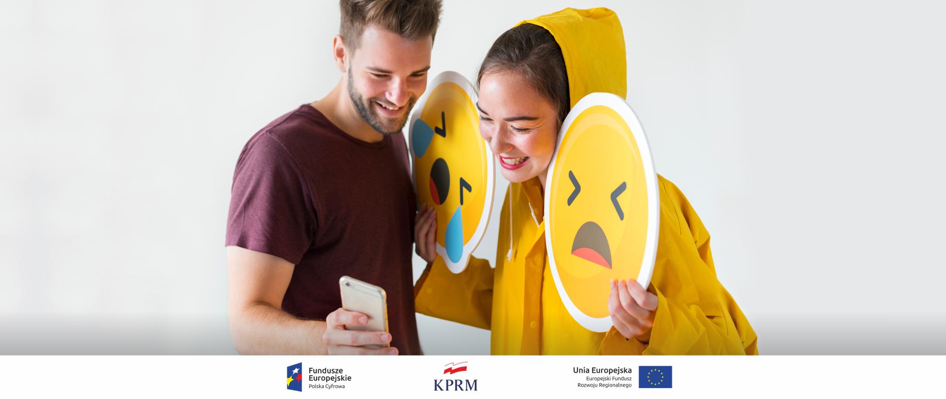 Dwie młode osoby kobieta i mężczyzna. Kobieta w żółtym sztormiaku trzyma w dłoniach (dwa) żółte emotikony (zapłakana buzia i smutna buzia). Uśmiechnięta kobieta patrzy w smartfoma trzymanego przez mężczyznę.