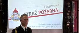 Na zdjęciu znajduje się strażak w mundurze, w tle widać slajd z prezentacji