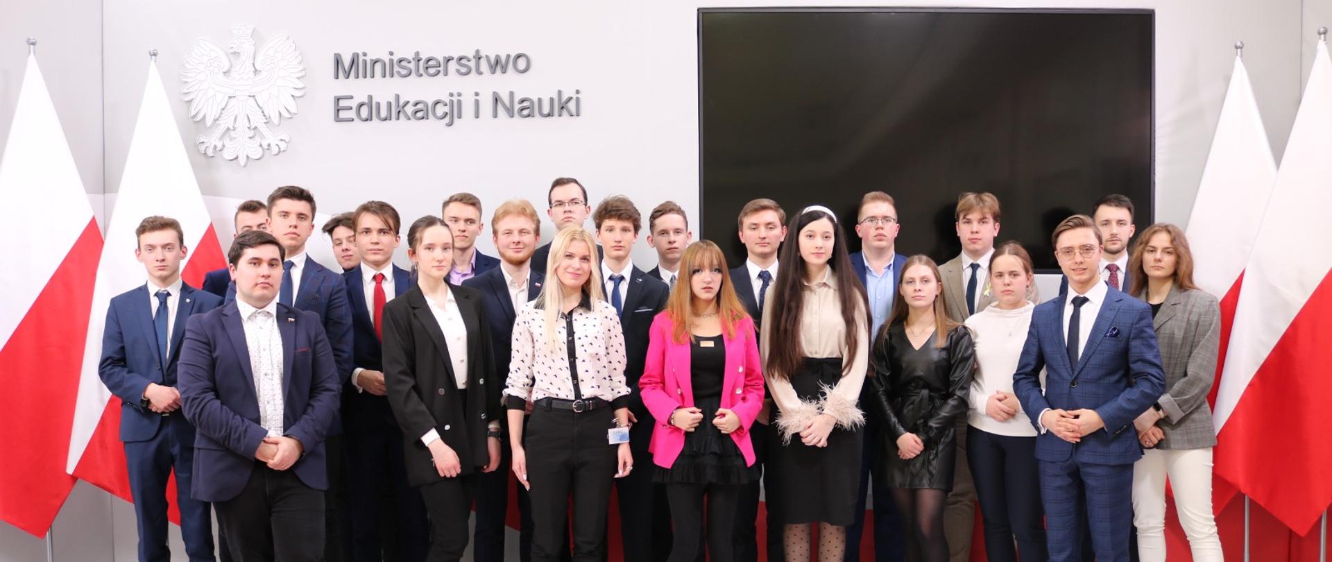 Grupa młodych ludzi, po środku dziewczyna ubrana w różowy żakiet. Po bokach biało-czerwone flagi. W tle oreł i napis Ministerstwo Edukacji i Nauki oraz duży ekran. 