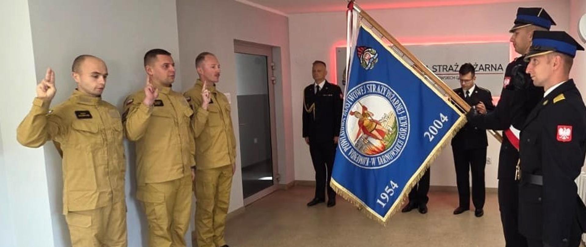 Na zdjęciu widzimy uroczystość ślubowania strażackiego.
Po lewej stronie stoją trzej mężczyźni w beżowych mundurach, unoszący prawą rękę z dwoma palcami w górze – co jest tradycyjnym gestem podczas składania przysięgi.
Po prawej stronie widzimy poczet sztandarowy – dwóch strażaków w czarnych, galowych mundurach trzyma sztandar jednostki straży pożarnej. Na sztandarze widnieje emblemat straży pożarnej, lata "1954 – 2004" oraz napis w języku polskim. Na ramieniu jednego z mundurów widać naszywkę z flagą Polski.
W tle, w pomieszczeniu, stoi kilku innych mężczyzn w mundurach galowych, obserwujących uroczystość. Na ścianie widnieje napis "STRAŻ POŻARNA".
Całość wygląda na oficjalną ceremonię w straży pożarnej, prawdopodobnie związana z przyjęciem nowych strażaków do służby.