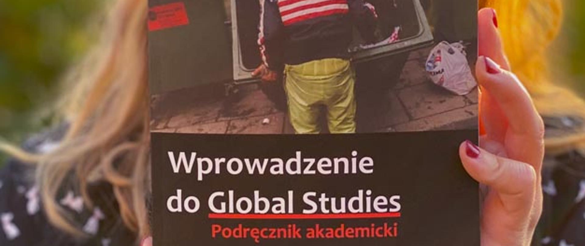 Na pierwszym planie okładka podręcznika akademickiego trzymanego w rękach przez kobietę