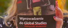 Na pierwszym planie okładka podręcznika akademickiego trzymanego w rękach przez kobietę