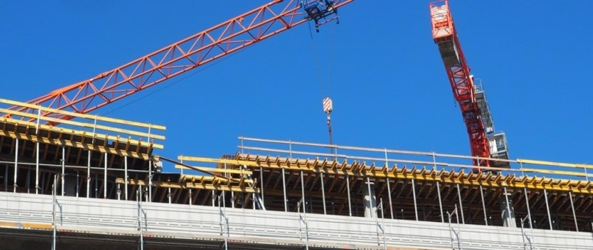 Dwa dźwigi pracujące na placu budowy – jeden poziomy pomarańczowy, drugi czerwony ustawiony pod kątem – unoszą materiały na konstrukcję budynku otoczoną rusztowaniami.