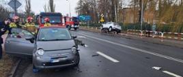 Zdjęcie przedstawia:
z lewe strony szaty samochód osobowy z otwartymi drzwiami po prawej stronie z uszkodzonym lewym bokiem. Obok stoi policjant i strażacy.
Po prawej stronie biały samochód osobowy po uderzeniu w barierki. Na ulicy stoją dwa samochody strażackie. 