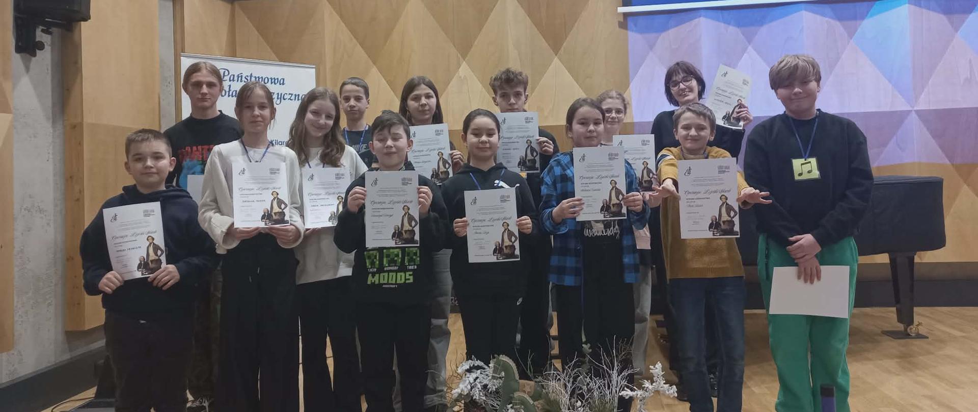 Uczniowie na scenie sali koncertowej z dyplomami uczestnictwa w wydarzeniu