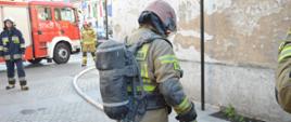 Pożar kamienicy na rawickim rynku. Na pierwszym planie strażak, który przed chwilą wyszedł z budynku objętego pożarem. Cały jest ubrudzony. Na plecach ma aparat powietrzny. W tle wóz strażacki, dwóch innych strażaków oraz fragment ratusza.