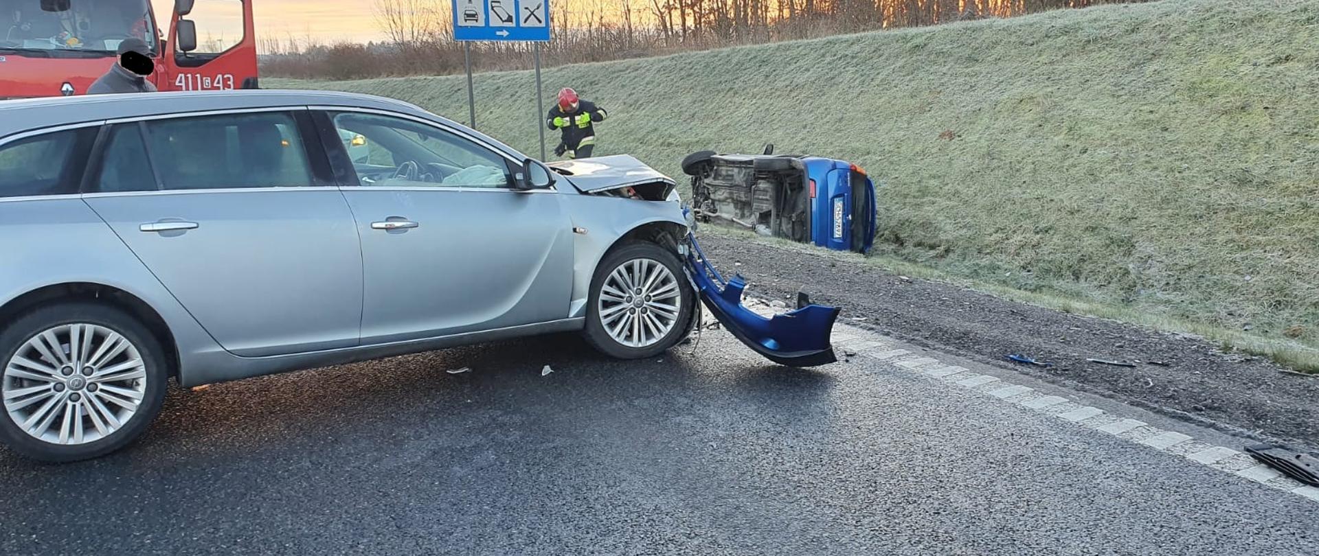 Samochód osobowy widziany z boku, stoi w poprzek na jezdni. Widoczne uszkodzenia przedniej części pojazdu. W tle za samochodem stoi wóz strażacki i widoczny jest drugi pojazd leżący w przydrożnym rowie. 