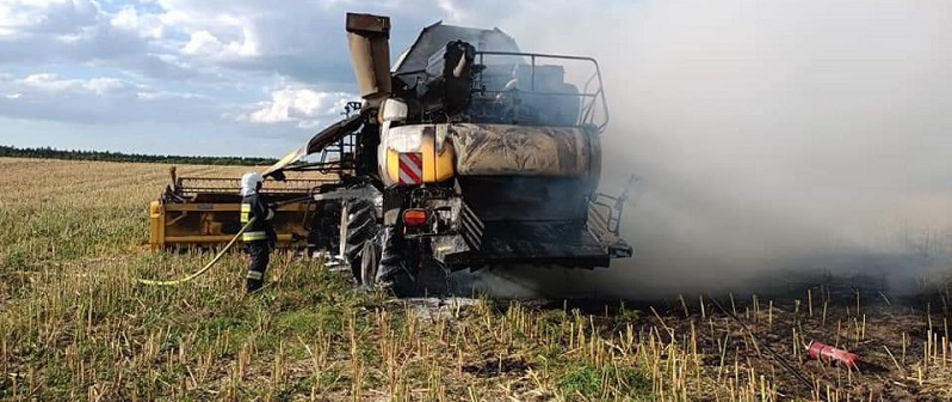 Przedstawia kombajn stojący na polu w którym doszło do pożaru. Obok strażak gaszący linią wodną palący się pojazd