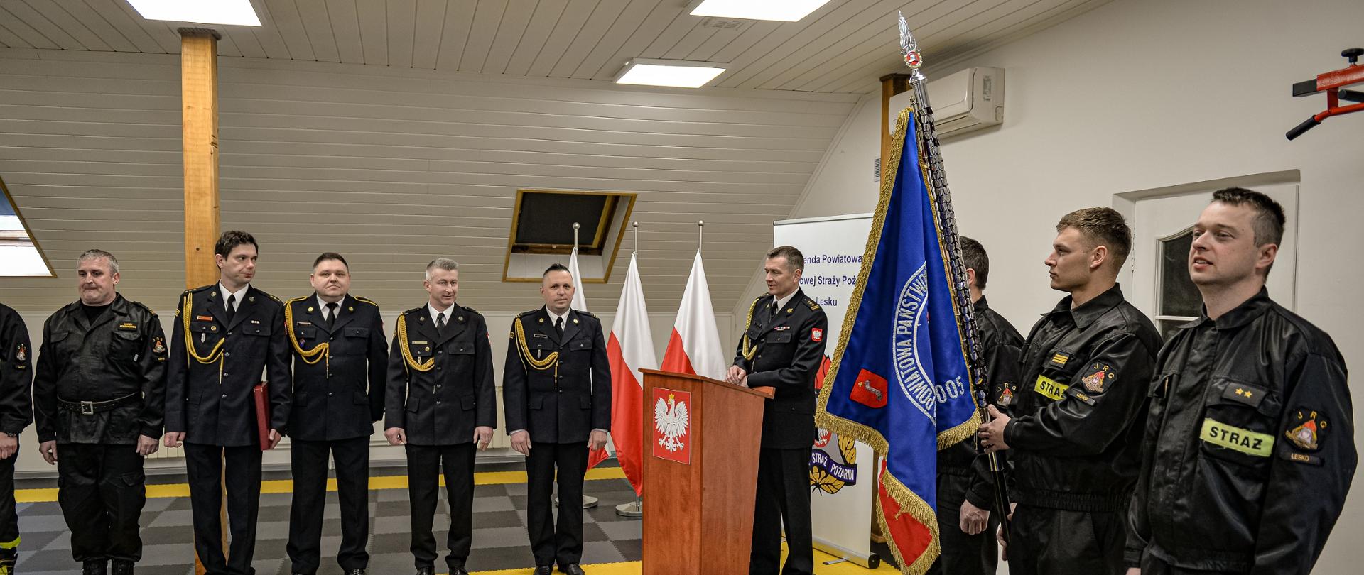 Zdjęcie przedstawia strażaków Komendy Powiatowej Państwowej Straży Pożarnej w Lesku podczas zbiórki z okazji przejścia na zaopatrzenie emerytalne Zastępcy Dowódcy Jednostki Ratowniczo – Gaśniczej w Lesku. W centralnej części kadru stoi trzech strażaków, jeden z nich trzyma sztandar Komendy Powiatowej Państwowej Straży Pożarnej w Lesku. Z lewej strony zdjęcia w jednym szeregu stoją strażacy w mundurach wyjściowych. 
