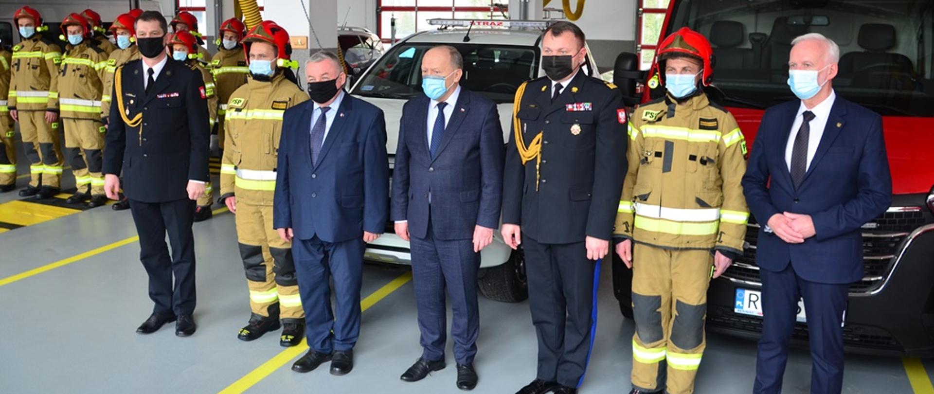 Mężczyźni stoją w postawie zasadniczej w szeregu na garażu przed dwoma samochodami pożarniczymi. Dwóch z nich występuje w umundurowaniu wyjściowym, dwóch w umundurowaniu bojowym, a pozostali w ubraniach cywilnych. W głębi widoczni strażacy w umundurowaniu bojowym, którzy stoją w dwuszeregu. 