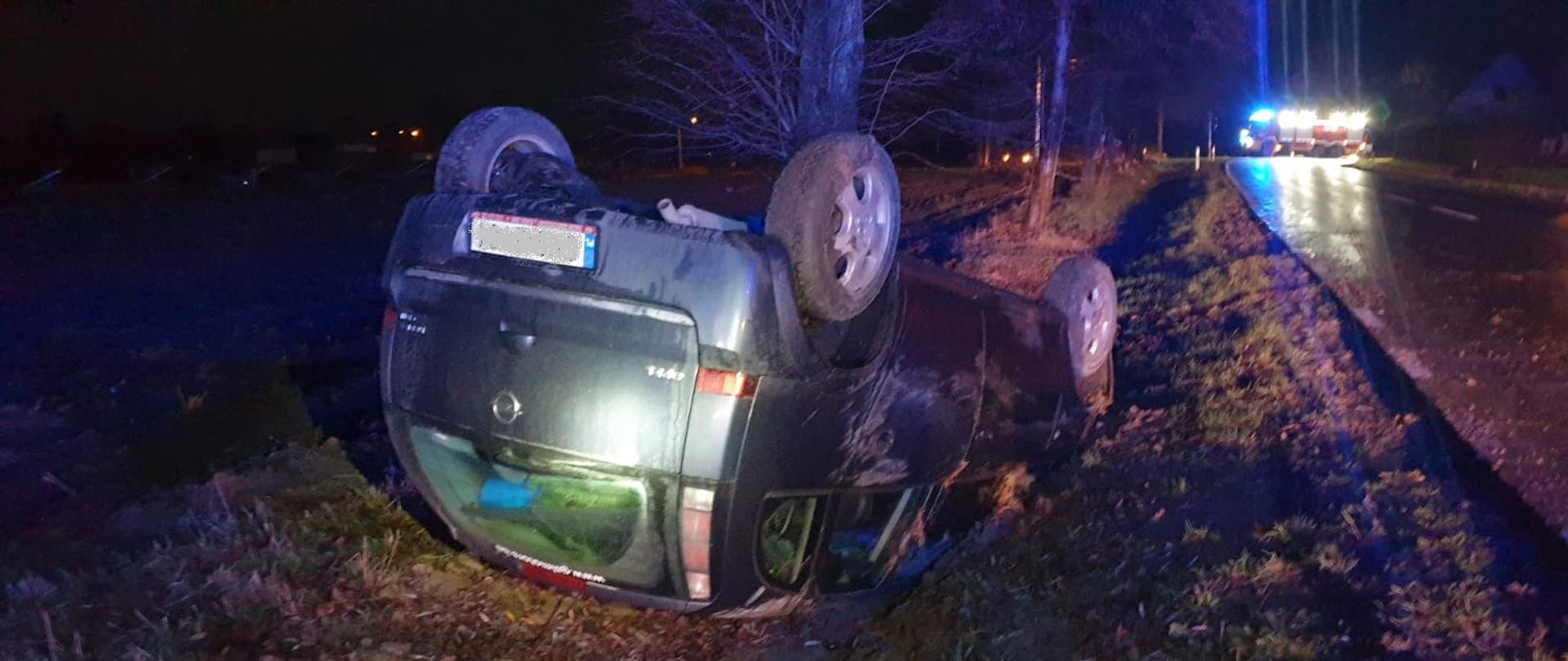 Po środku samochód osobowy Opel leży na dachu w przydrożnym rowie. W oddali na drodze stoi oświetlony samochód straży pożarnej