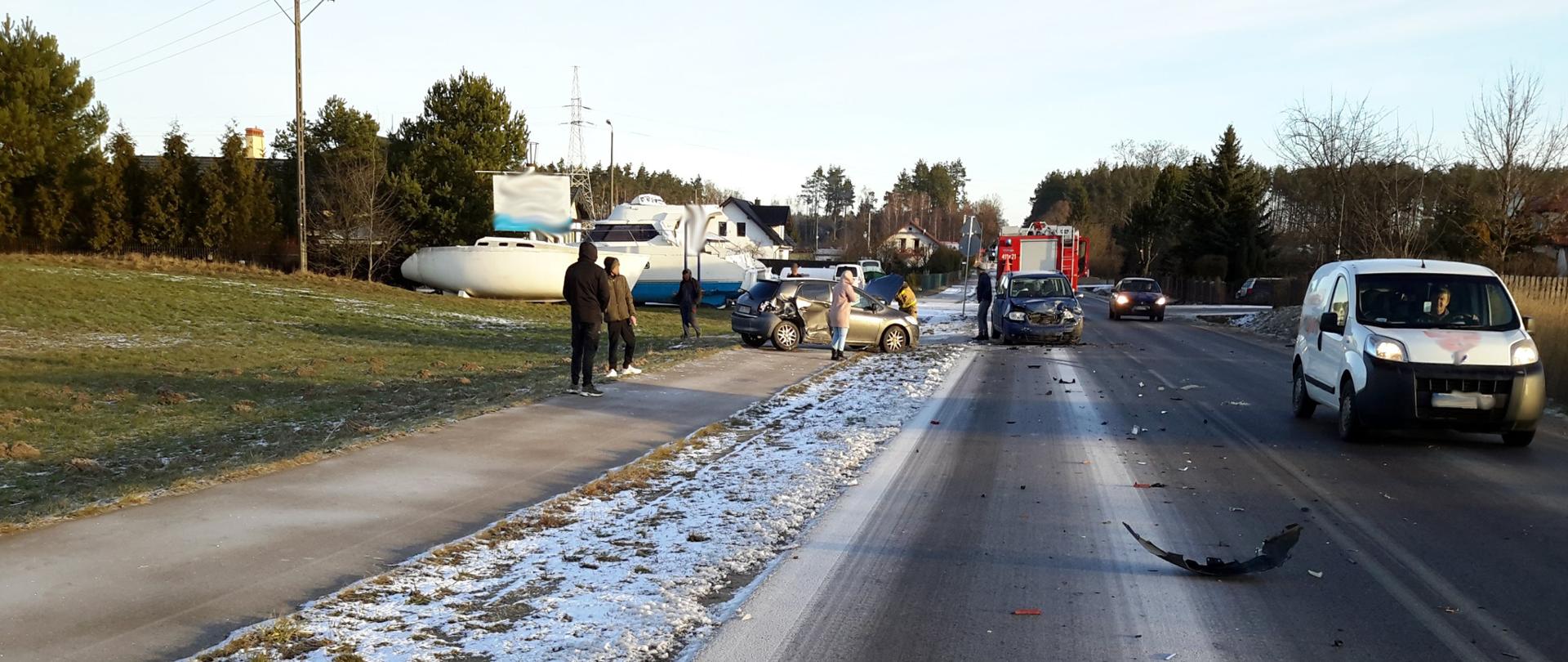 Samochód osobowy z rozbitym przodem i bokiem stoi na poboczu drogi na chodniku. Strażak zabezpiecza samochód. W tle osoby postronne, inny samochód uczestniczący w kolizji, wóz strażacki oraz kadłuby łodzi z pobliskiego zakładu szkutniczego. Na drodze rozrzucone części samochodowe.