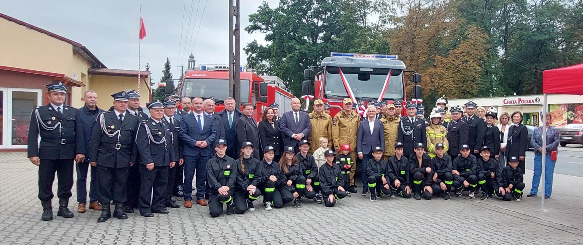 Strażacy i osoby cywilne stoją w kilku szeregach na tle samochodów strażackich.