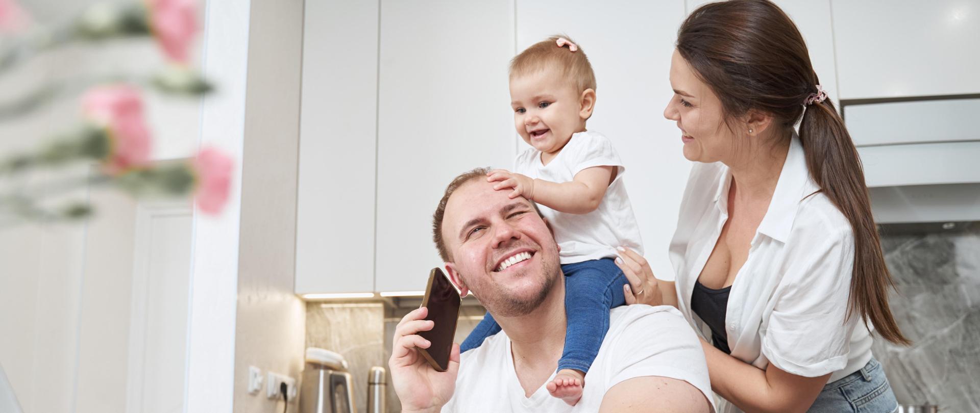 Wife holding baby on neck of her husband, while he working with laptop and answering phone call