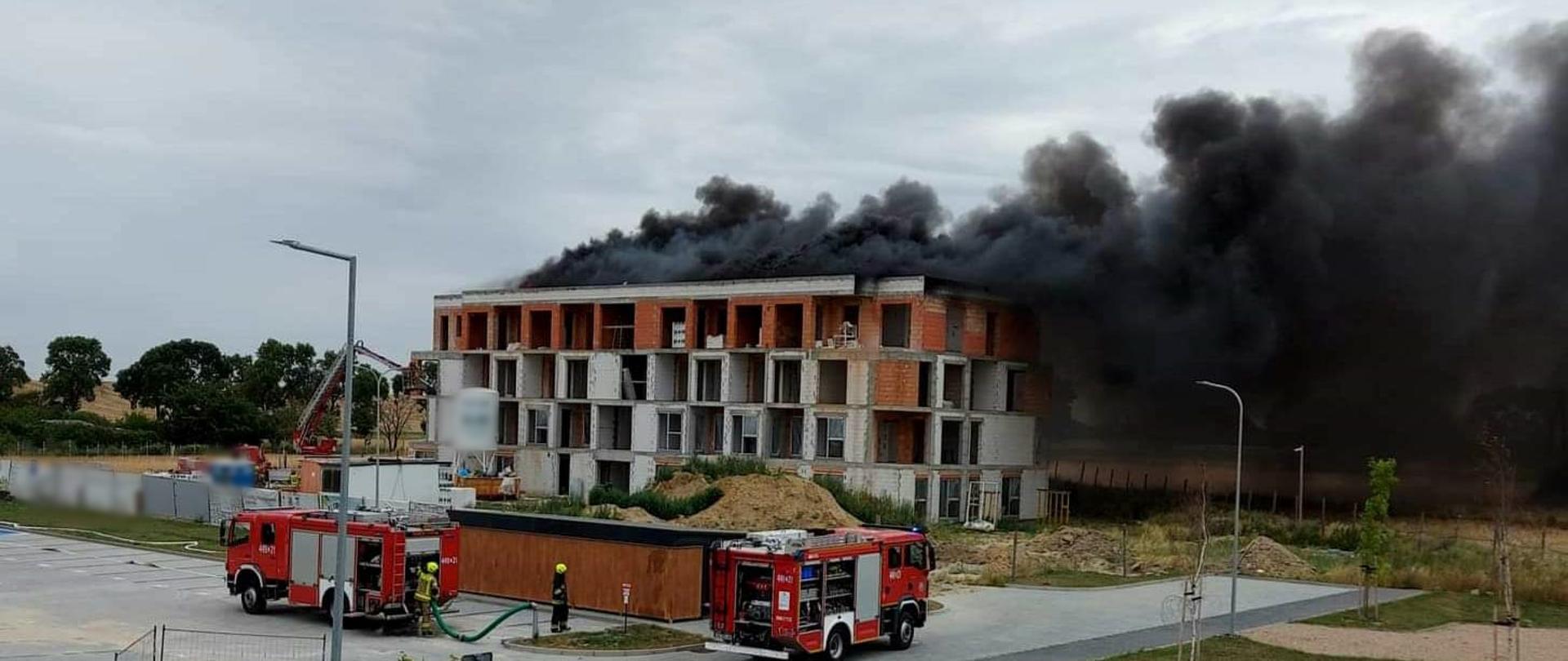 Obraz przedstawia płonący dach budowanego budynku wielorodzinnego. Widoczny czarny dym. Przed budynkiem pojazdy pożarnicze. Pora dzienna.