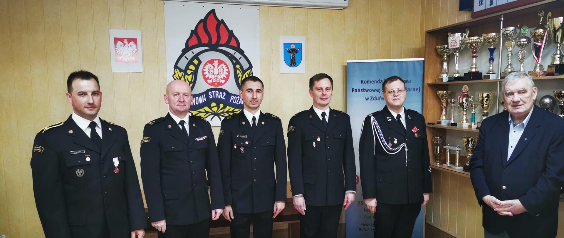 Na zdjęciu widoczni odznaczeni funkcjonariusze PSP w umundurowaniu wyjściowym zlokalizowani w pomieszczeniu. Na ścianie w tle widoczne duże logo PSP oraz roll-up Komendy Powiatowej PSP w Zduńskiej Woli. 