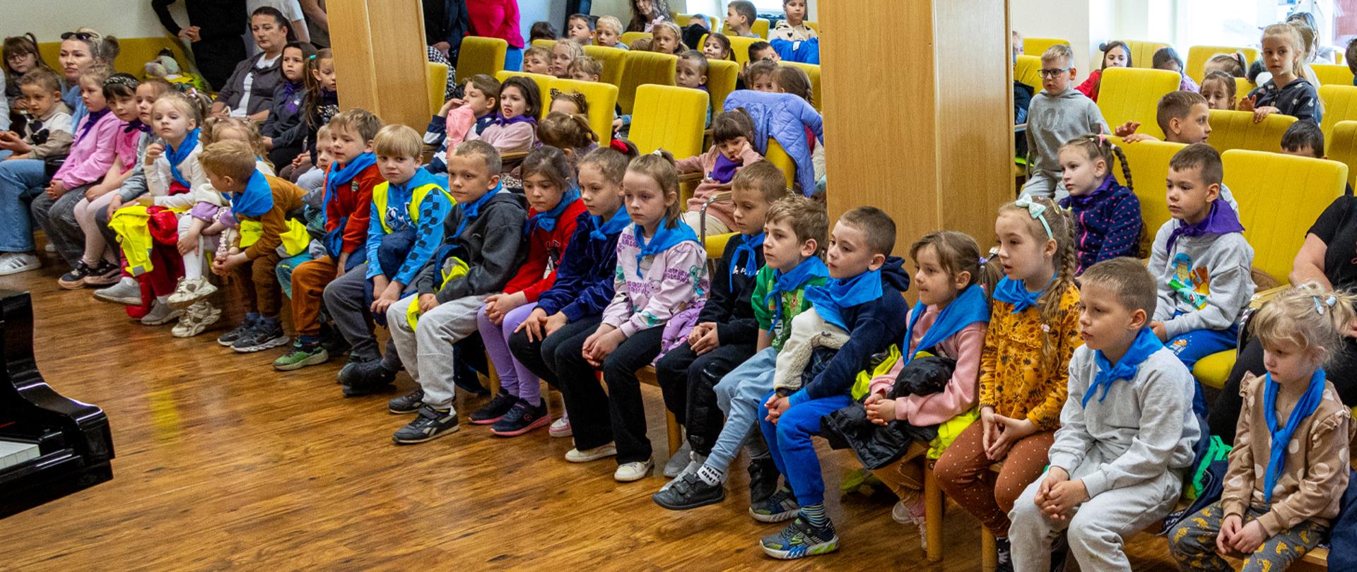 Na zdjęciu duża grupa przedszkolaków wraz z wychowawczyniami siedzi w auli szkolnej.