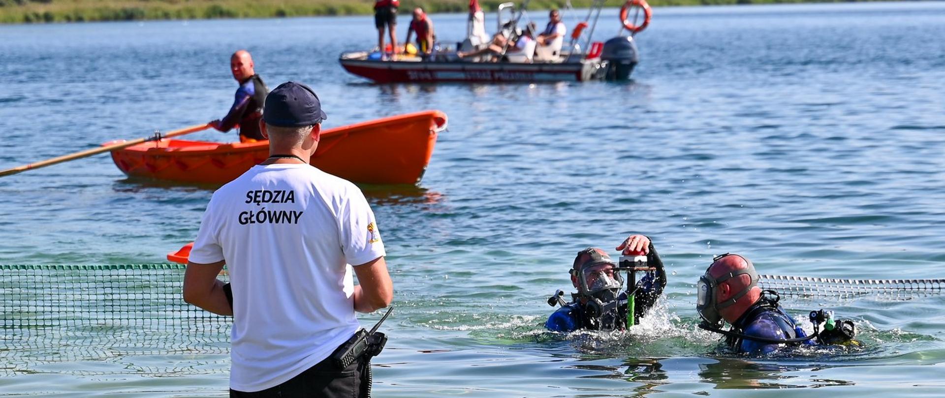 Dwóch nurków w pełnym sprzęcie wynurza się przy siatce w wodzie przed sędzią głównym zawodów. Jeden z nurków naciska przycisk kończąc konkurencję.który obserwuje konkurencję.