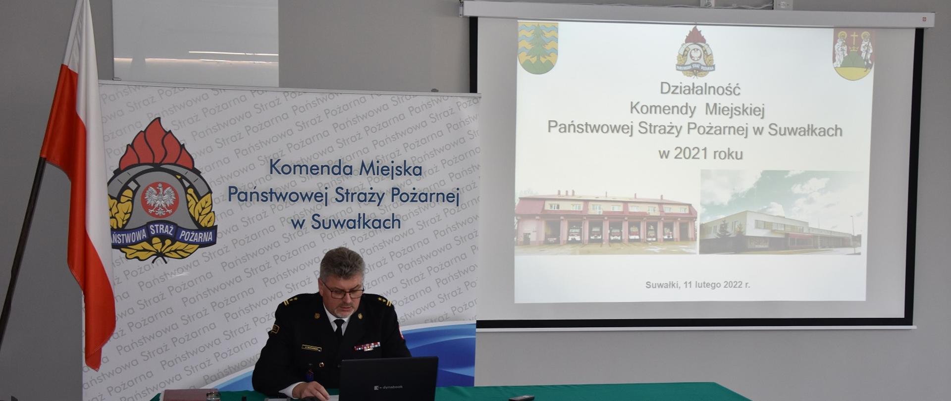 Zdjęcie obrazuje strażaka za biurkiem przy komputerze w tle flaga RP logo PSP z napisem Komenda Miejska Państwowej Straży Pożarnej w Suwałkach i planszą z prezentacją dotycząca działalności jednostek ochrony przeciwpożarowej powiatu suwalskiego w 2021 r.