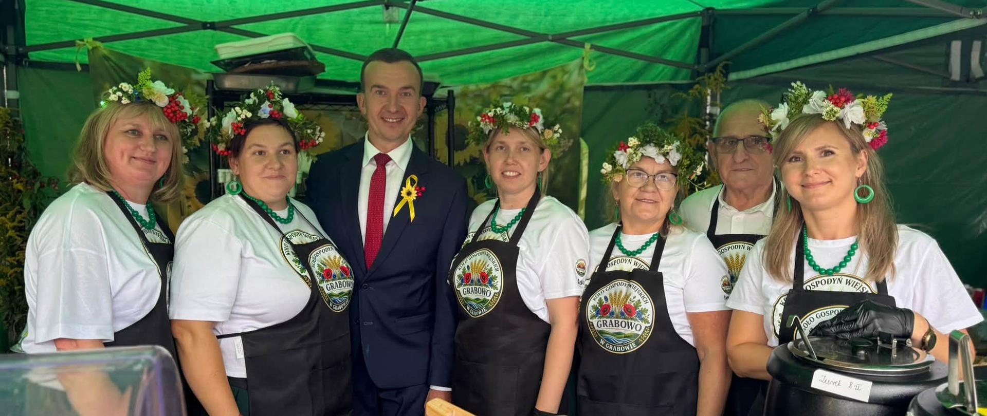 Mężczyzna w granatowym garniturze stoi pod zielonym namiotem wśród kobiet w jednakowych fartuszkach i wiankach z kwiatów.
