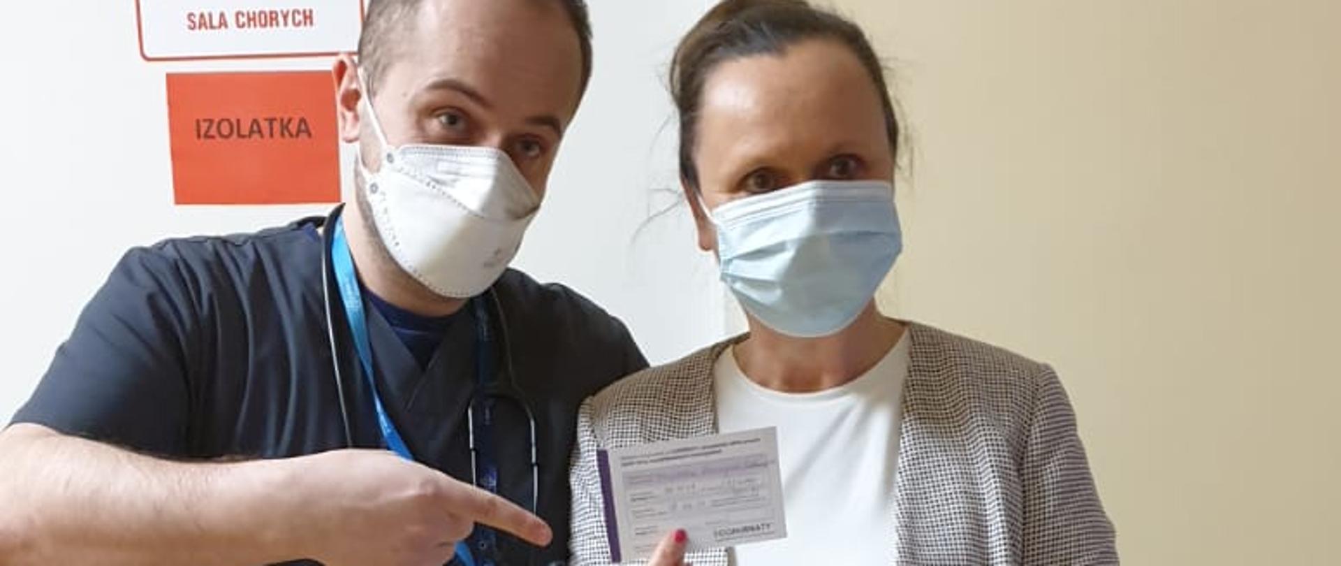Kobieta w koku (ciemne włosy) średniego wzrostu w szarej spódnicy i marynarce w biało-czarną kratkę. Obok mężczyzna ze stetoskopem w granatowym T-shircie i ciemnych spodniach.
Mężczyzna unosi palec wskazujący wskazując na zaświadczenie trzymane przez kobietę o przebytym szczepieniu.
