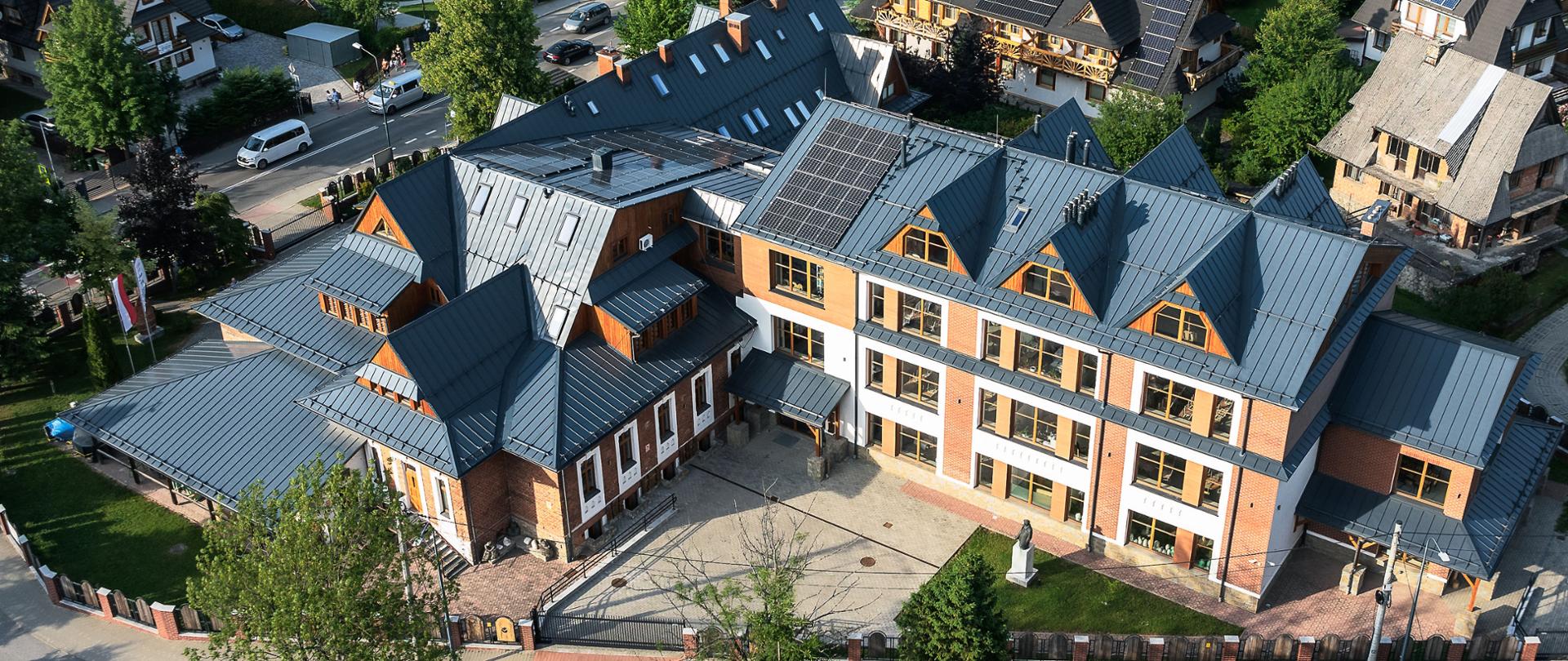 Na zdjęciu widzimy budynek głównej siedziby szkoły „STRUG” przy ul. Kościeliskiej 35 w Zakopanem. Fotografia wykonana jest przy użyciu drona i całość budynku widzimy z lotu ptaka. Widzimy z szarym spadzistym dachem, ściany budynku wykonane są z czerwonej cegły. Okna budynku w kolorze brązowym mają opasane są białymi ramami wykonanymi z tynku. Wokół budynku widzimy sąsiednie domy, ulice oraz roślinność i tatrzański pejzaż z górami.