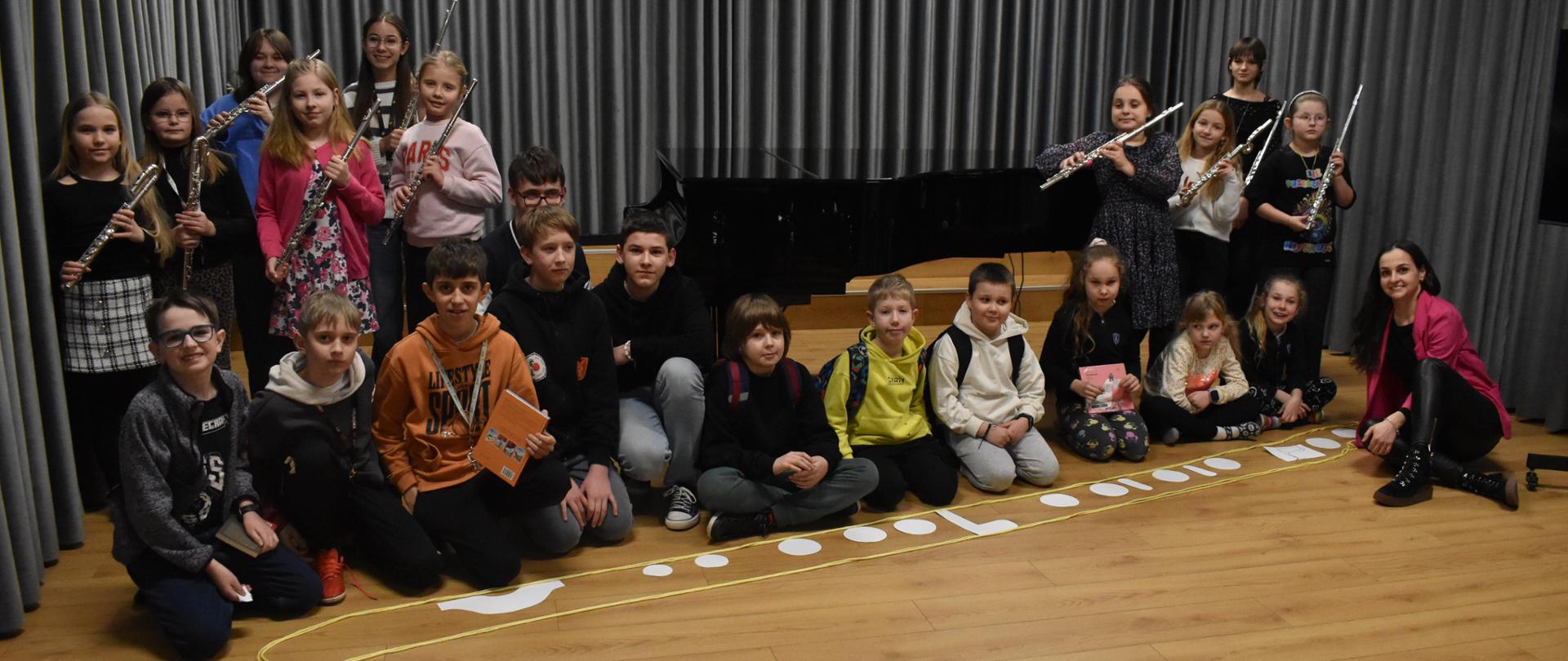 na zdjęciu, w auli PSM widnieją uczniowie i nauczycielka, którzy stworzyli na scenie największy flet świata ze sznurka i materiałów papierniczych. Część uczniów trzyma w ręku flety, a niektórzy nagrody książkowe. Kolorystyka zdjęcia jest biało-szaro-brązowo-czerwono-żółto-niebiesko-czarna. 