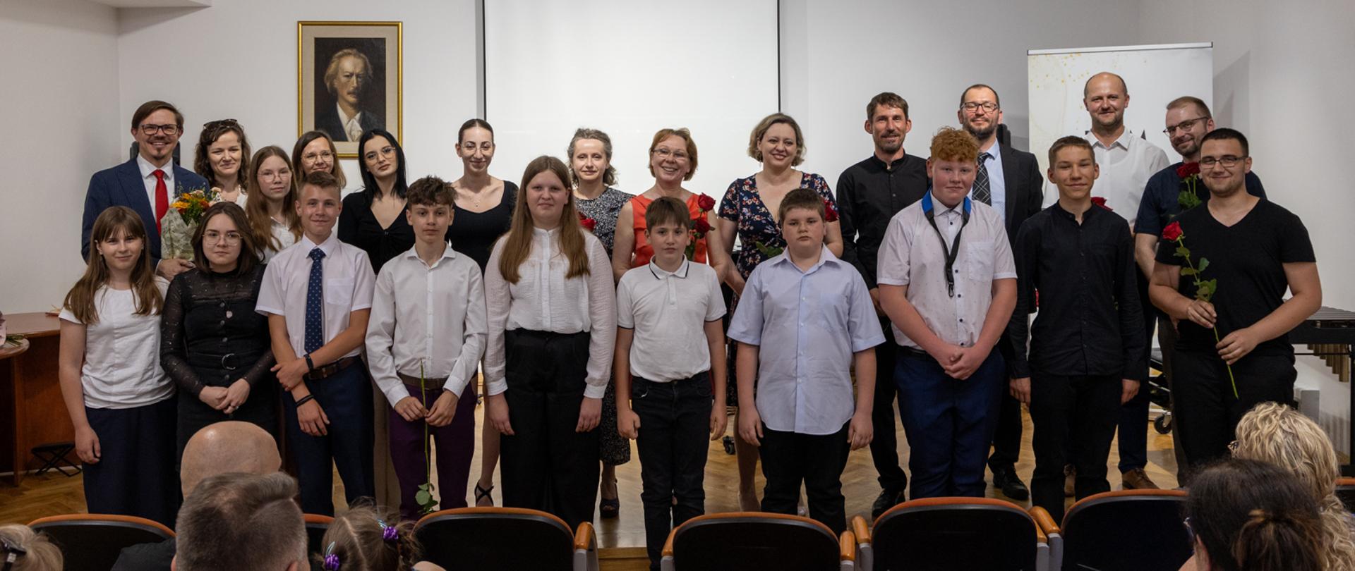 Zdjęcie grupowe przedstawiające absolwentów roku szkolnego 2022/2023 wraz z ich nauczycielami. Nauczyciele stoją na scenie PSM w Głubczycach, absolwenci przed stopniem stanowiącym granicę sceny. Na dole zdjęcia widać również kilka osób siedzących na publiczności.