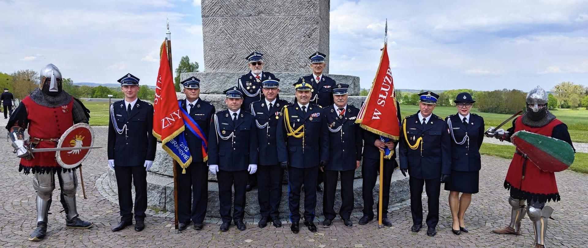 Na zdjęciu przy pomniku stoi grupa strażaków OSP w mundurach galowych i dwa sztandary. Na środku Warmińsko mazurski komendant wojewódzki PSP. Po bokach dwie osoby przebrane za rycerzy.
