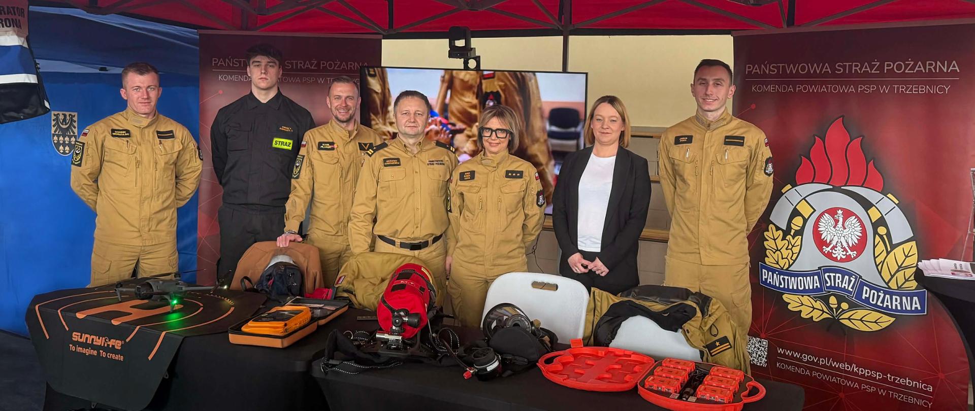 Strażacy pod namiotem z napisem Państwowa Straż Pożarna. Przed nimi na stole sprzęt strażacki. 