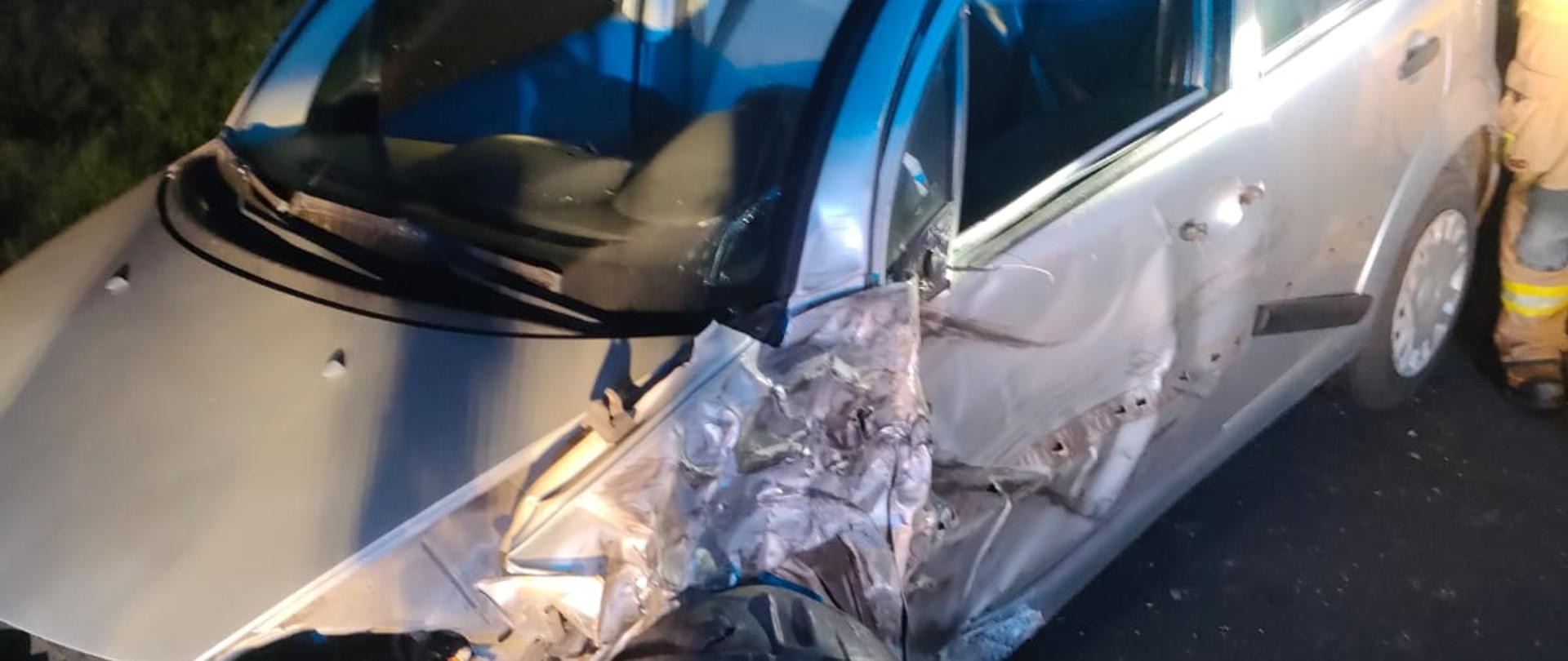 Na zdjęciu widać srebrny samochód osobowy stojący na drodze nocą. Pojazd ma poważnie uszkodzony przód i lewą stronę, szczególnie w rejonie koła oraz drzwi kierowcy. W tle widoczne są światła służb ratunkowych, a obok samochodu stoi strażak w umundurowaniu.