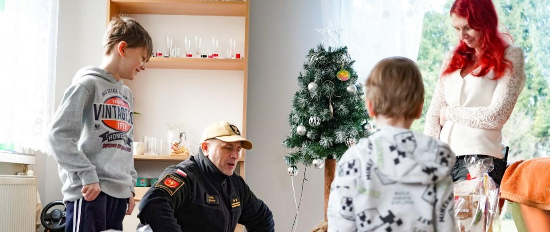 Zdjęcie przedstawia komendanta powiatowego oraz kobietę i dwóch chłopców. Rodzina odbiera prezenty od komendanta. Komendant kuca. 