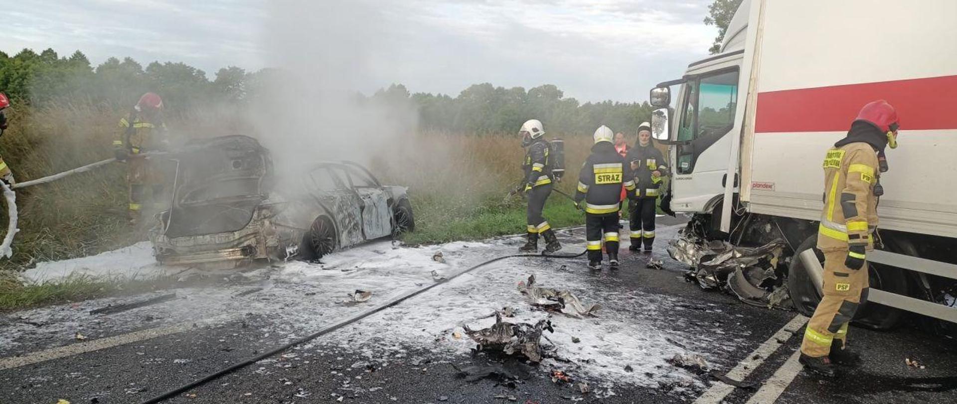 Zdjęcie wykonane w dzień. Na drodze stoi rozbite, tlące się jeszcze auto. Przy aucie strażacy w aparatach ODO. Trwa akcja ratunkowa.